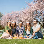Diverse group of friends at a picnic looking up with awe, all wearing cherry blossom design eclipse glasses under a sunny sky.