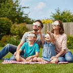 Family of four on a picnic blanket, all wearing Doodle solar eclipse glasses while looking up at the sky, CE certified.