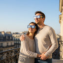 Pareja romántica en un balcón parisino, con gafas diseño EU, mirando el cielo despejado con asombro.