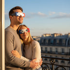 Romantic couple on a Paris balcony wearing FR eclipse glasses, looking at the sky with awe, perfect for August 12, 2026.