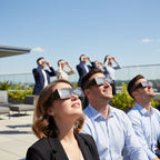 Colleagues at a corporate event wearing goldline eclipse glasses, captivated by the sky during a sunny day.