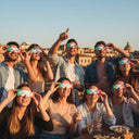 Freunde feiern auf einem römischen Dach, tragen Sonnenfinsternis-Brillen im IT-Design, teilen ein Lächeln und blicken zum Himmel.