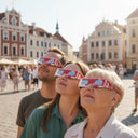 Grupo diverso en una reunión comunitaria en Letonia, todos con gafas para eclipse solar y mirando al cielo juntos.