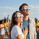 Ein glückliches junges Paar mit Neon-Sonnenfinsternis-Brille auf einer sonnendurchfluteten Dachterrasse, das gemeinsam zum hellen Himmel blickt.