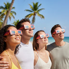 Four friends on a tropical beach, all wearing solar eclipse glasses and looking up at the bright sky in shared wonder.