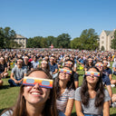A large crowd at a university event, all wearing peace design glasses, looking up with expressions of awe.