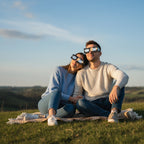 A young couple on a grassy hilltop, wearing solar eclipse glasses and gazing at the sky in awe. August 12, 2026.