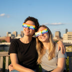 A loving couple wearing rainbow solar eclipse glasses, sharing a joyful moment on a sunlit rooftop garden.