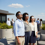 Colleagues at a wellness event on a rooftop terrace, all wearing reflective eclipse glasses and looking up with shared excitement.