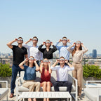 Seven friends at a rooftop party, all wearing Swing design eclipse glasses, sharing a moment of wonder under the bright sky.