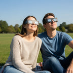 Happy couple wearing Swing design eclipse glasses, gazing at the sky in a tranquil park, ready for the August 12, 2026 event.