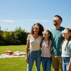 Group of friends in a backyard, wearing Wood design eclipse glasses, sharing smiles while looking up at the bright sky. ISO certified.